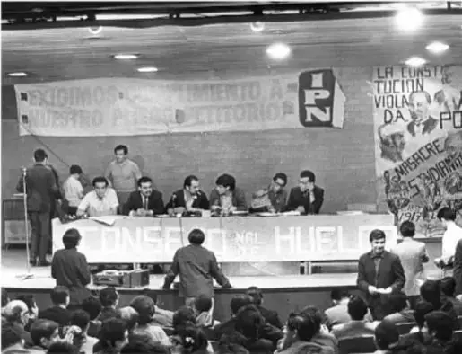 Fotografía en blanco y negro de una asamblea estudiantil en un auditorio. Se observan personas sentadas en una mesa larga frente a una audiencia, con mantas y carteles al fondo. Una manta dice '¡EXIGIMOS AUMENTO A NUESTRO PRESUPUESTO EDITORIAL!' y otra '¡CNH!'. También hay un cartel con la silueta de un puño y la palabra 'IPN'.