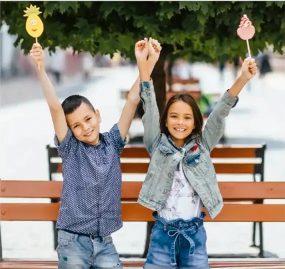 Fotografía de dos niños sentados en una banca de parque, con los brazos levantados sosteniendo dulces o paletas, en un ambiente alegre y soleado.