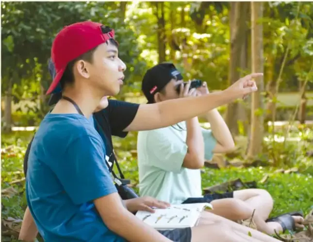 Dos jóvenes en un entorno natural; uno señala hacia arriba mientras el otro sostiene binoculares, observando aves o el dosel del bosque.