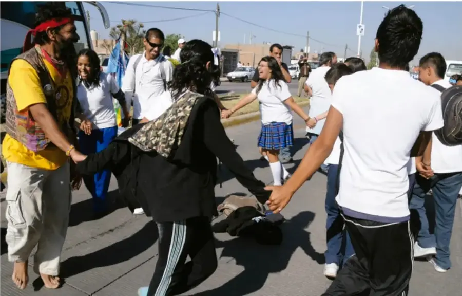 Fotografía: un grupo de jóvenes estudiantes con uniforme escolar y dos adultos (uno con chaleco amarillo y pañuelo rojo) forman un círculo tomados de las manos sobre la calle, en un espacio público soleado con autos y edificios al fondo; parece una dinámica o juego cooperativo.