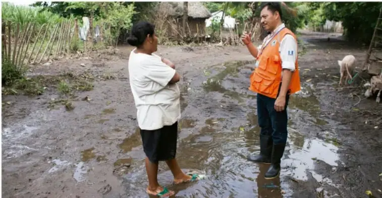 Fotografía de una entrevista en una comunidad rural: una mujer del lugar conversa con un entrevistador con chaleco naranja en un camino de tierra mojado, con casas de bahareque y vegetación al fondo; ilustra la actividad de entrevistar a personas mayores de la comunidad.
