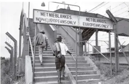 Fotografía en blanco y negro de una escalera con dos letreros: «NET NIE BLANKES / NON-EUROPEANS ONLY» y «NET BLANKES / EUROPEANS ONLY», separando el acceso por raza durante el apartheid.