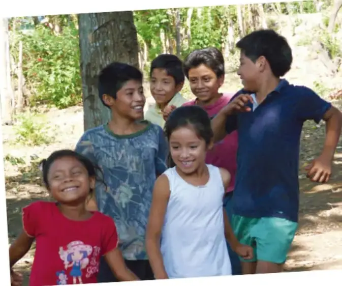 Fotografía de un grupo de niños y niñas sonriendo y jugando al aire libre, rodeados de árboles, lo que ilustra la convivencia y el juego comunitario.