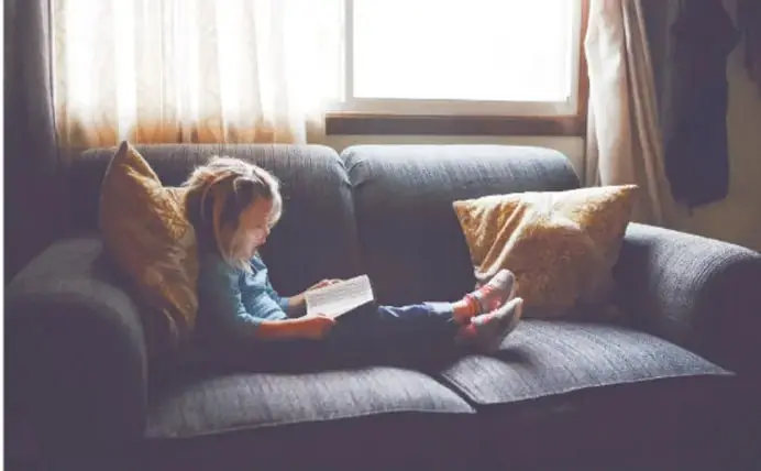 Niña leyendo un libro sentada en un sillón junto a una ventana.