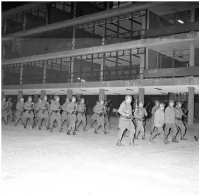Fotografía en blanco y negro de soldados o granaderos avanzando en formación por una calle, con equipo antidisturbios.