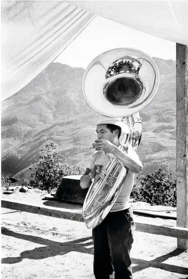 Fotografía en blanco y negro de un joven tocando un instrumento de viento (trombón/tuba) en un espacio abierto, con montañas de la sierra zapoteca al fondo.
