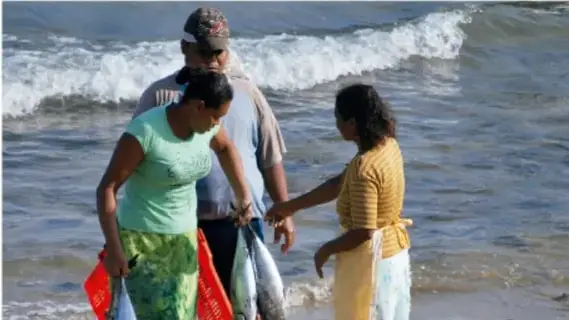Tres personas afrodescendientes conversan en la orilla de una playa junto a las olas, con redes y bolsas de pesca.