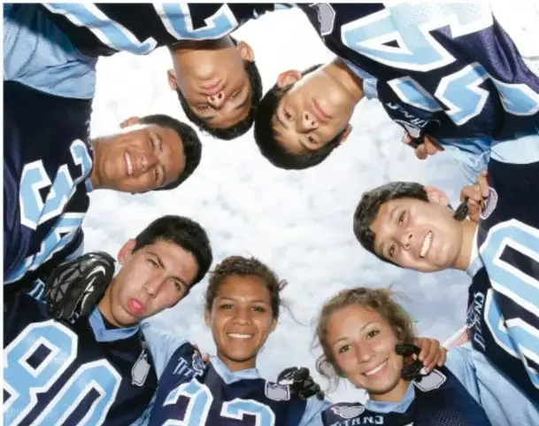 Fotografía de un grupo de estudiantes (adolescentes) con camisetas deportivas, mirando hacia arriba en círculo, sonriendo; ilustra el trabajo en comunidad.