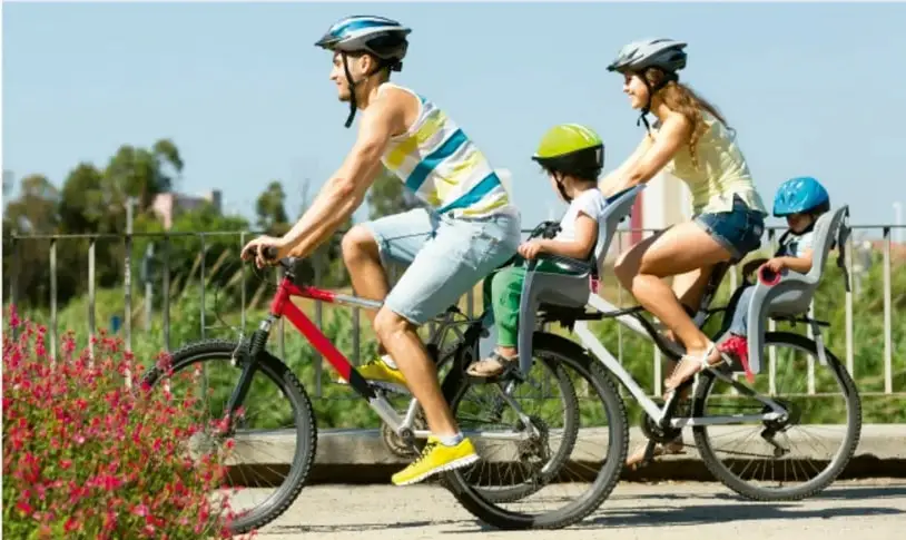 Fotografía de una familia (dos adultos y dos niños con casco) montando bicicletas en un parque con flores rojas y vegetación, ilustrando la actividad física familiar y el estilo de vida activo.