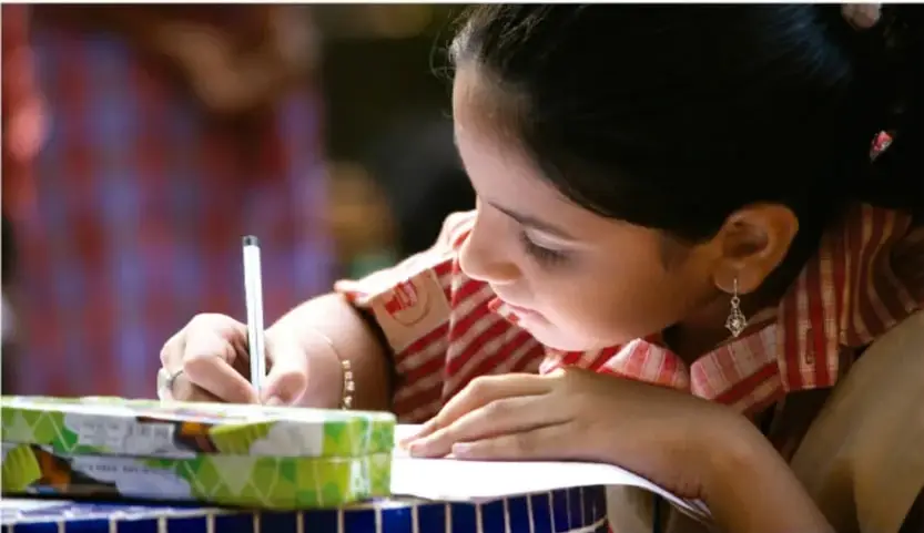 Fotografía de una niña escribiendo con un lápiz sobre una hoja, apoyada en una mesa con mosaicos azules; al fondo se ve a otra persona con camisa a cuadros.