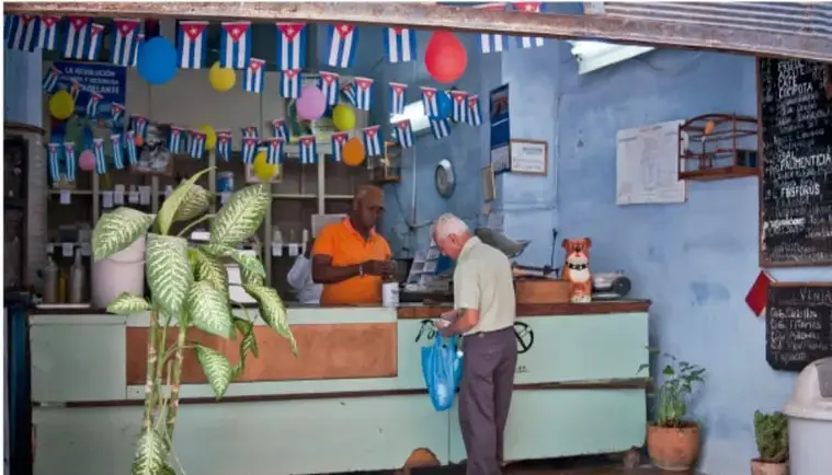 Local comercial en Cuba decorado con banderines y globos, con un cliente y un vendedor, ilustrando la actividad económica y la vida cotidiana en una zona poblada del Caribe.