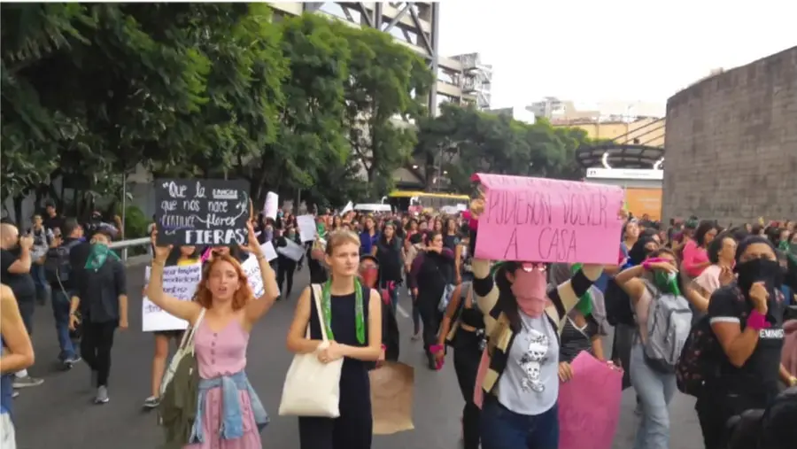 Fotografía de una marcha en la vía pública con personas que portan pancartas y pañuelos; una pancarta rosa dice 'PUEDEN VOLVER A CASA' y otra negra menciona 'Que la tierra que nos vio nacer nunca florezca sin nosotras'.