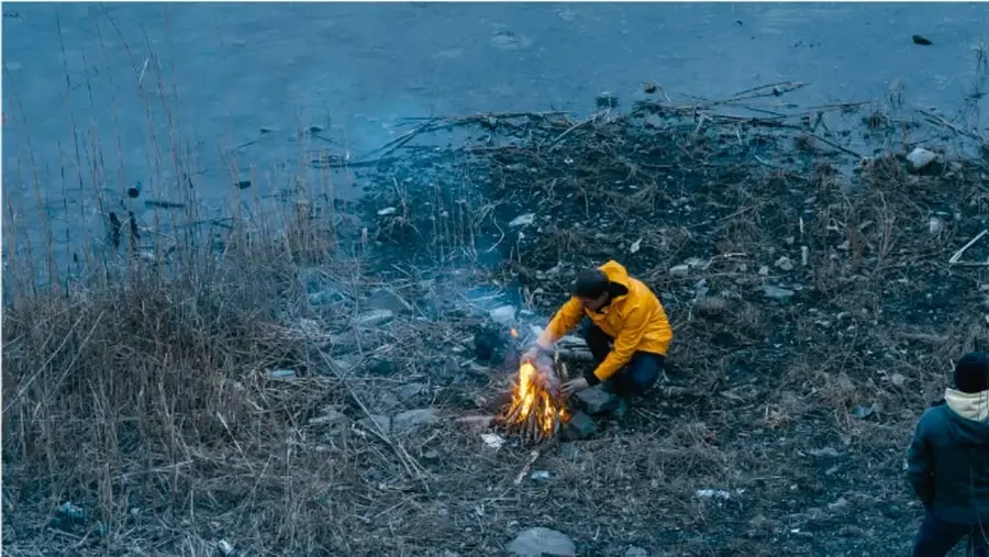 Fotografía de una persona con chamarra amarilla encendiendo una fogata en un terreno con basura y pasto seco cerca de un río, con otra persona al costado; ilustra el deterioro del ambiente.