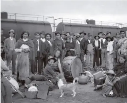 Fotografía histórica en blanco y negro de un grupo de personas (hombres, mujeres y niños) con sombreros y ropa de época, de pie junto a un vagón de tren, con un perro pequeño en primer plano; ilustra la sociedad mexicana del periodo revolucionario.