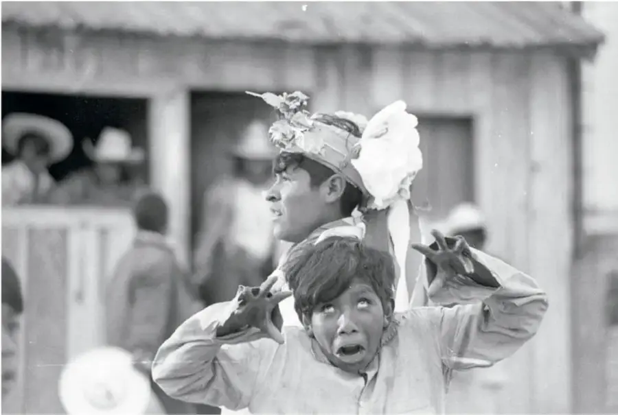Fotografía en blanco y negro de una construcción de madera con una mujer asomada a la puerta; al fondo, personas reunidas. Título: Tepehuanos, semana santa, 1978, Héctor García (1923-2012).