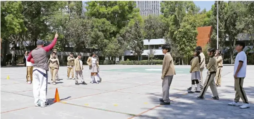 Fotografía de estudiantes de secundaria en uniforme en una cancha escolar al aire libre, con un docente que levanta el brazo dando indicaciones; hay conos naranjas y líneas amarillas marcadas en el piso, edificaciones y árboles al fondo.