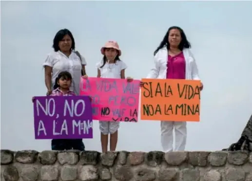 Fotografía de cuatro personas (tres mujeres y una niña) de pie sobre un muro de piedra, sosteniendo carteles con lemas en contra de la minería: 'NO, NO A LA MINA', 'LA VIDA... NO A LA MINA'.