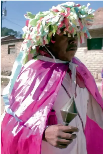 Fotografía de un hombre indígena ataviado con un tocado de papeles de colores (rosa, verde, blanco) y capa rosa, sosteniendo un objeto ceremonial; ilustra las ceremonias de los pueblos originarios descritas en el texto.