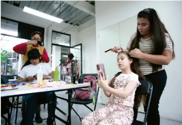 Fotografía de un taller comunitario: una joven peina a una niña mientras otras personas realizan actividades manuales en mesas compartidas, ilustrando redes de apoyo y ambientes saludables.