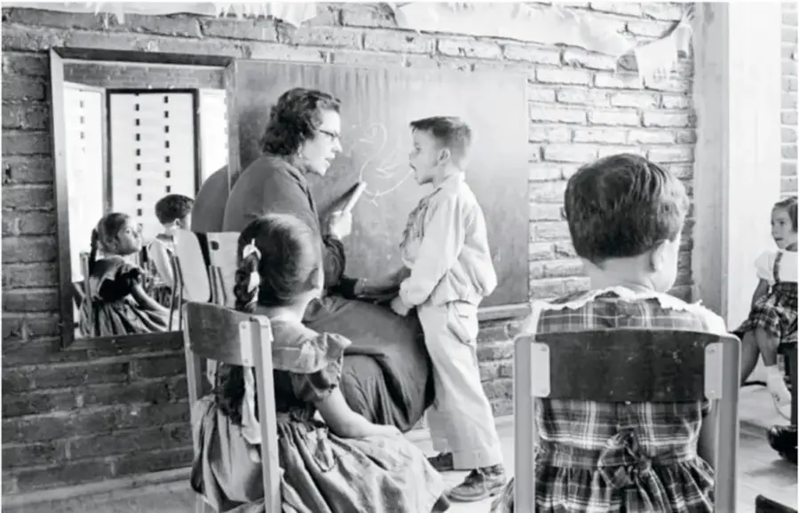 Clase al aire libre: una maestra con un niño frente al pizarrón junto a una pared de ladrillo, niñas sentadas en sillas escolares y un reflejo de otras niñas en un marco.