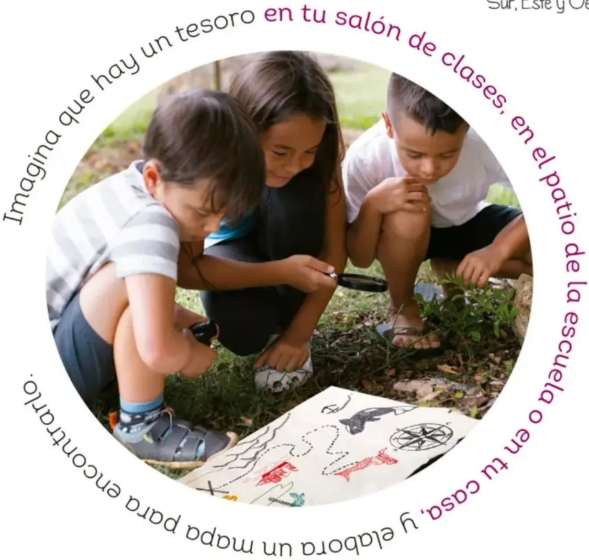 Tres niños revisando con lupa un mapa del tesoro dibujado a mano sobre el pasto; el enunciado de la actividad corre en círculo alrededor de la fotografía.
