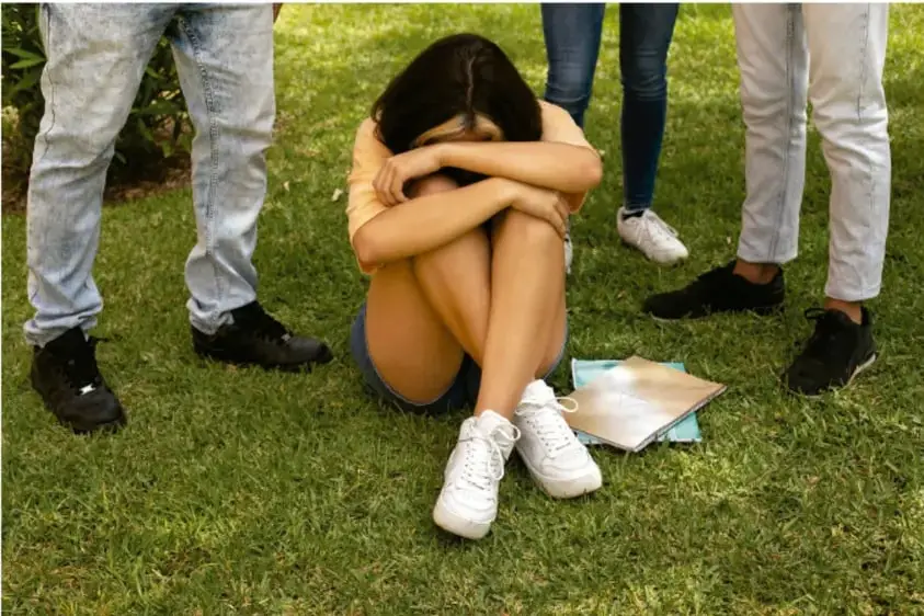 Fotografía de una joven sentada en el pasto con la cabeza entre las rodeada por personas de pie; junto a ella hay un libro y una máscara sanitaria en el suelo.