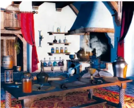 Fotografía de un laboratorio o taller de alquimista con matraces, frascos de vidrio de colores, un crisol con fuego sobre una mesa de madera, y una chimenea al fondo.