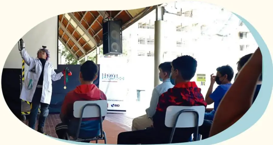 Estudiantes observan una obra de teatro escolar en un patio con techo de lámina.