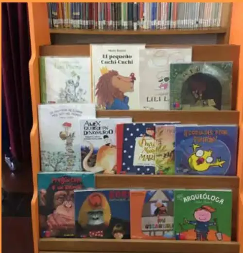 Fotografía de una biblioteca de aula con estantería de madera naranja y libros infantiles exhibidos de frente, como 'El pequeño Cuchi Cuchi', 'Alicia en el país de las maravillas' y 'Miquelóloga'. Pie de foto: 'Foto 2. La Biblioteca de aula debe estar accesible a los estudiantes.'