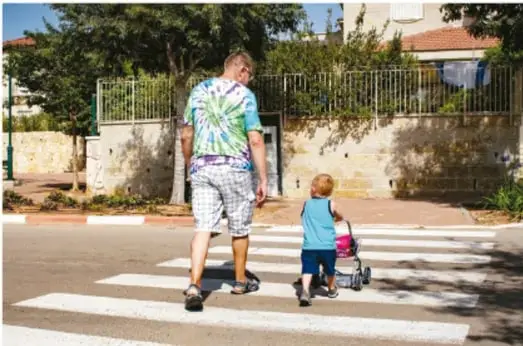 Adulto cruzando un paso de peatones acompañado de una niña pequeña que empuja un cochecito, imagen que ejemplifica ir acompañado de una persona adulta por seguridad.