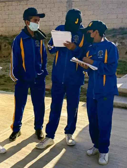 Fotografía de tres estudiantes de telesecundaria con uniforme azul y amarillo, mascarillas, sosteniendo cuadernos y pluma, tomada al aire libre; crédito: Luis Eduardo García Medel, Tlaxiaca.