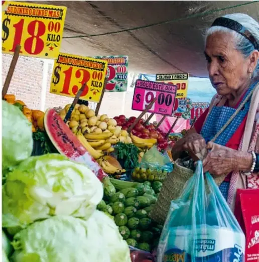 Mujer mayor comprando verduras en un mercado; carteles de precios visibles: $18.00 el kilo, $13.00 el kilo y $9.00, junto a cabbage, sandía y otros productos.