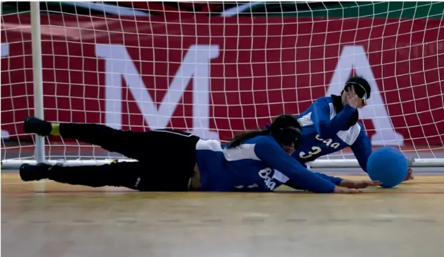 Fotografía de jugadoras de goalball en pista cubierta: una atleta en el suelo extendida hacia el balón azul, ambas con antifaces, camisetas azules, y la portería con fondo rojo con letras blancas al fondo.