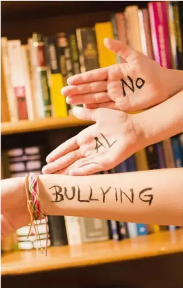 Fotografía de manos con las palabras 'No AL BULLYING' escritas en la piel, frente a un estante con libros.