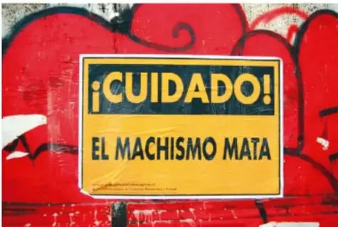Fotografía de un muro con grafiti rojo y un cartel amarillo con letras negras que dice '¡CUIDADO! EL MACHISMO MATA'.