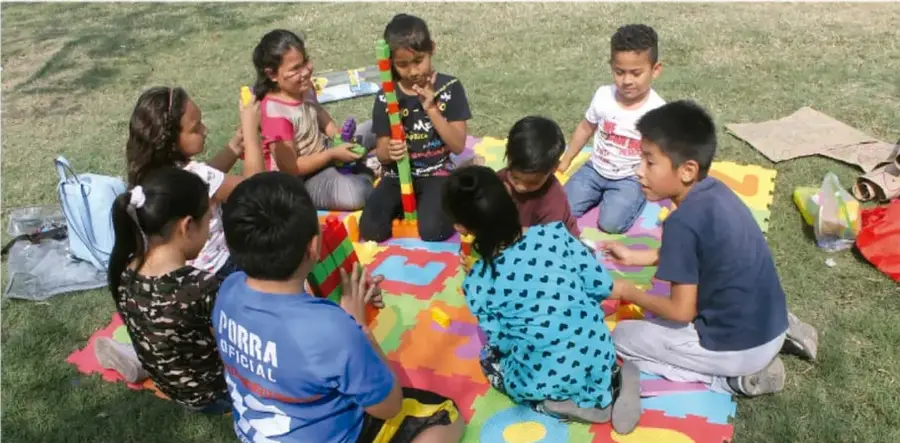 Grupo de niñas y niños sentados en el pasto sobre un tapete de colores, jugando con bloques y materiales didácticos.
