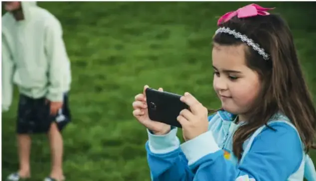 Una niña con diadema rosa y chamarra azul toma una fotografía con un teléfono celular; al fondo, un niño de espaldas camina en un parque con pasto verde.