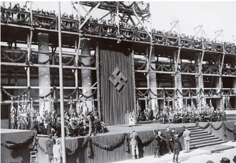 Fotografía en blanco y negro de un gran desfile o concentración masiva frente a un edificio con columnas y una gran bandera con la esvástica nazi.