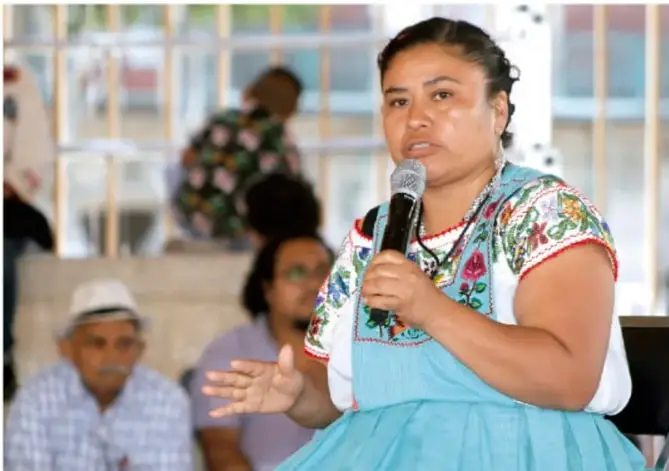 Mujer indígena con vestimenta tradicional bordada toma la palabra con un micrófono durante una asamblea comunitaria, con otros miembros de la comunidad sentados de fondo.