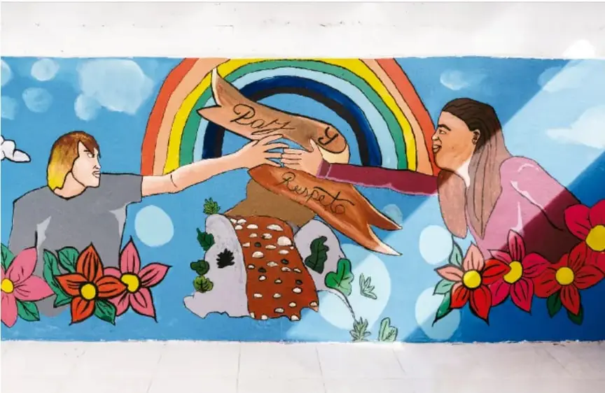 Fotografía de un mural en el que dos jóvenes estrechan las manos frente a un arcoíris; un pergamino entre sus manos dice "Paz y respeto"; hay flores rojas y follaje verde en la parte inferior y un cielo azul con nubes de fondo.