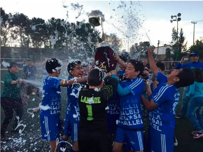 Fotografía de un grupo de niños de un equipo de fútbol celebrando con un trofeo, rociándose con espuma o agua, en actitud de alegría compartida.