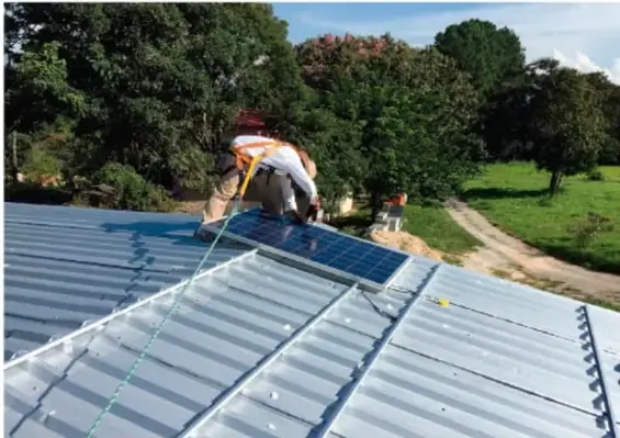 Persona trabajando sobre el techo de una casa instalando paneles solares, rodeada de árboles y campo.