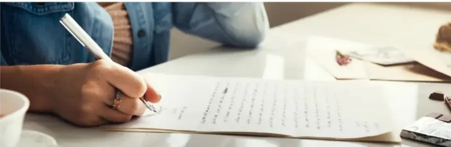 Fotografía de una persona escribiendo a mano una carta en una mesa, con pluma, taza de café, sobres y tarjeta postal, acompañada de un florero pequeño.