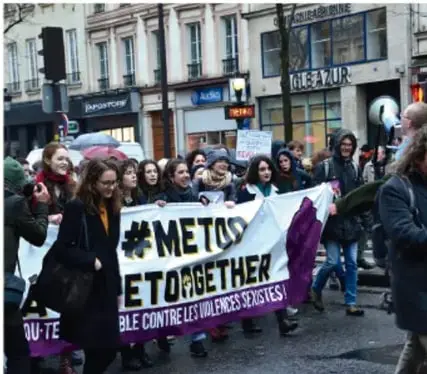 Fotografía de una marcha en la vía pública donde mujeres sostienen una pancarta con la leyenda '#MeToo ET TOGETHER' y 'LUTTE CONTRE LES VIOLENCES SEXISTES', en un contexto urbano con edificios y transeúntes.