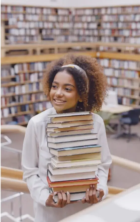 Fotografía de una estudiante sonriente con uniforme escolar que sostiene una pila alta de libros frente a los estantes de una biblioteca.