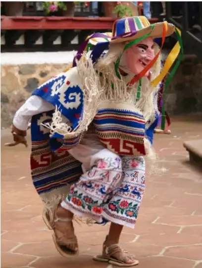 Persona disfrazada con traje tradicional colorido, sombrero y máscara, danzando en un patio empedrado, probablemente durante una fiesta comunitaria.