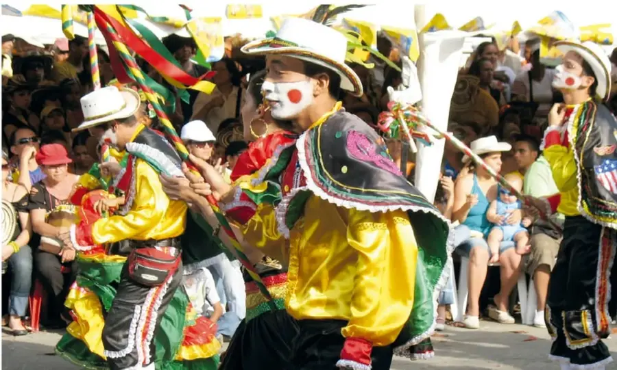 Grupo de danzantes con rostro pintado de blanco con círculos rojos, sombrero blanco, capa bordada y camisa amarilla bailando en la calle.