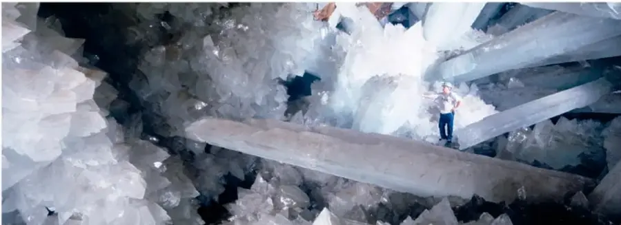 Fotografía de la Cueva de los Cristales de Naica con enormes vigas de cristales de yeso; una persona de pie sobre uno de ellos sirve de escala.