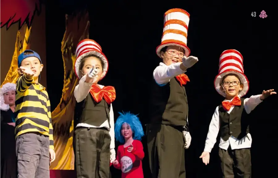 Fotografía de una obra escolar infantil: cuatro niños con sombreros altos rojos y blancos, corbatines naranjas y chalecos, en poses teatrales; una niña con peluca azul y playera roja, ante telones con árboles pintados.