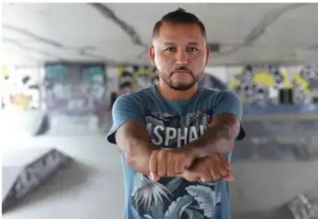 Fotografía de Pedro Carrizales, el Mijis, hombre joven con barba y cabello corto, usando una playera azul con la palabra 'SPHA'. Está de pie con los brazos cruzados en un espacio con grafitis.